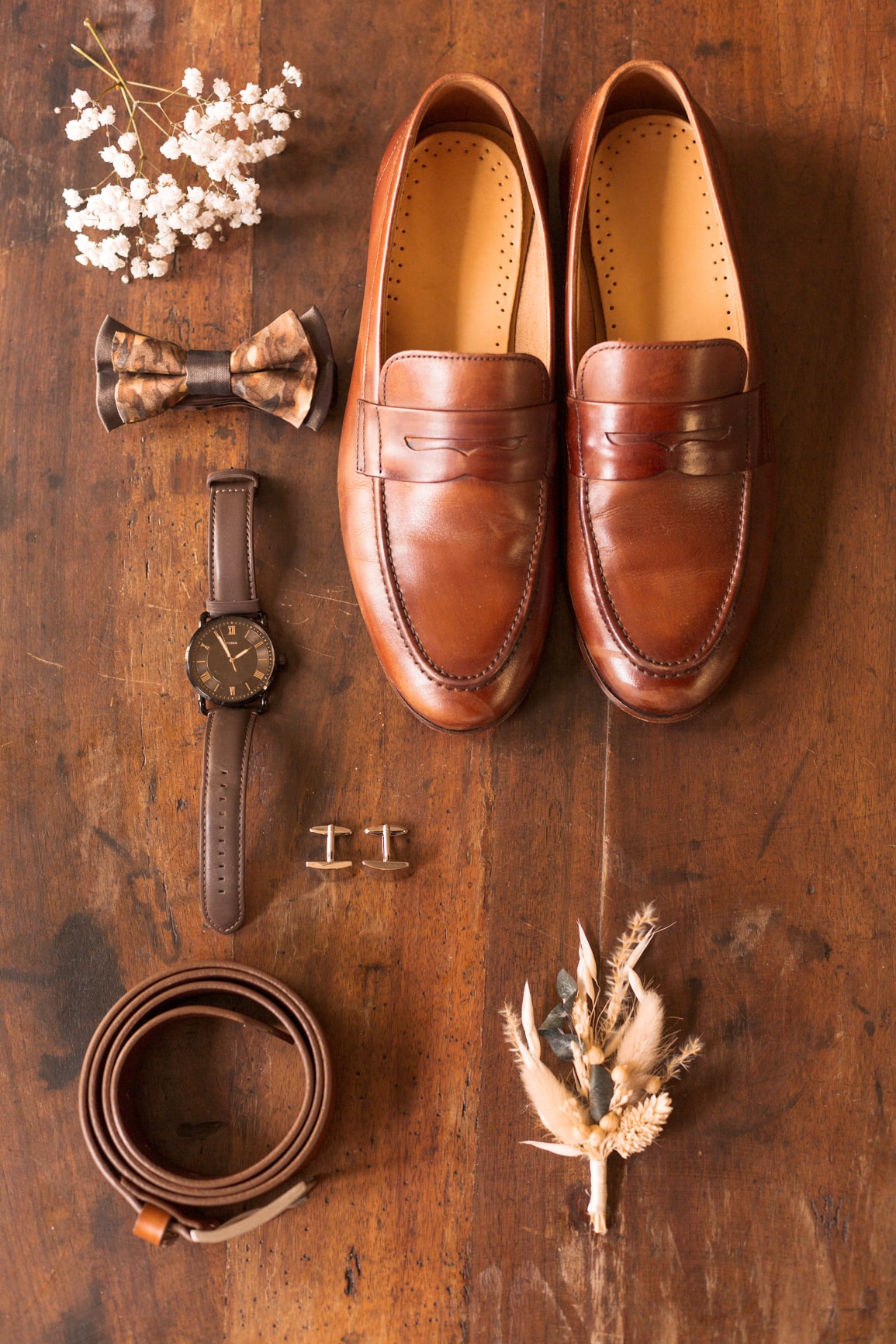 Accessoires du marié posés sur une table en bois : chaussures en cuir marron, montre, ceinture et boutonnière.