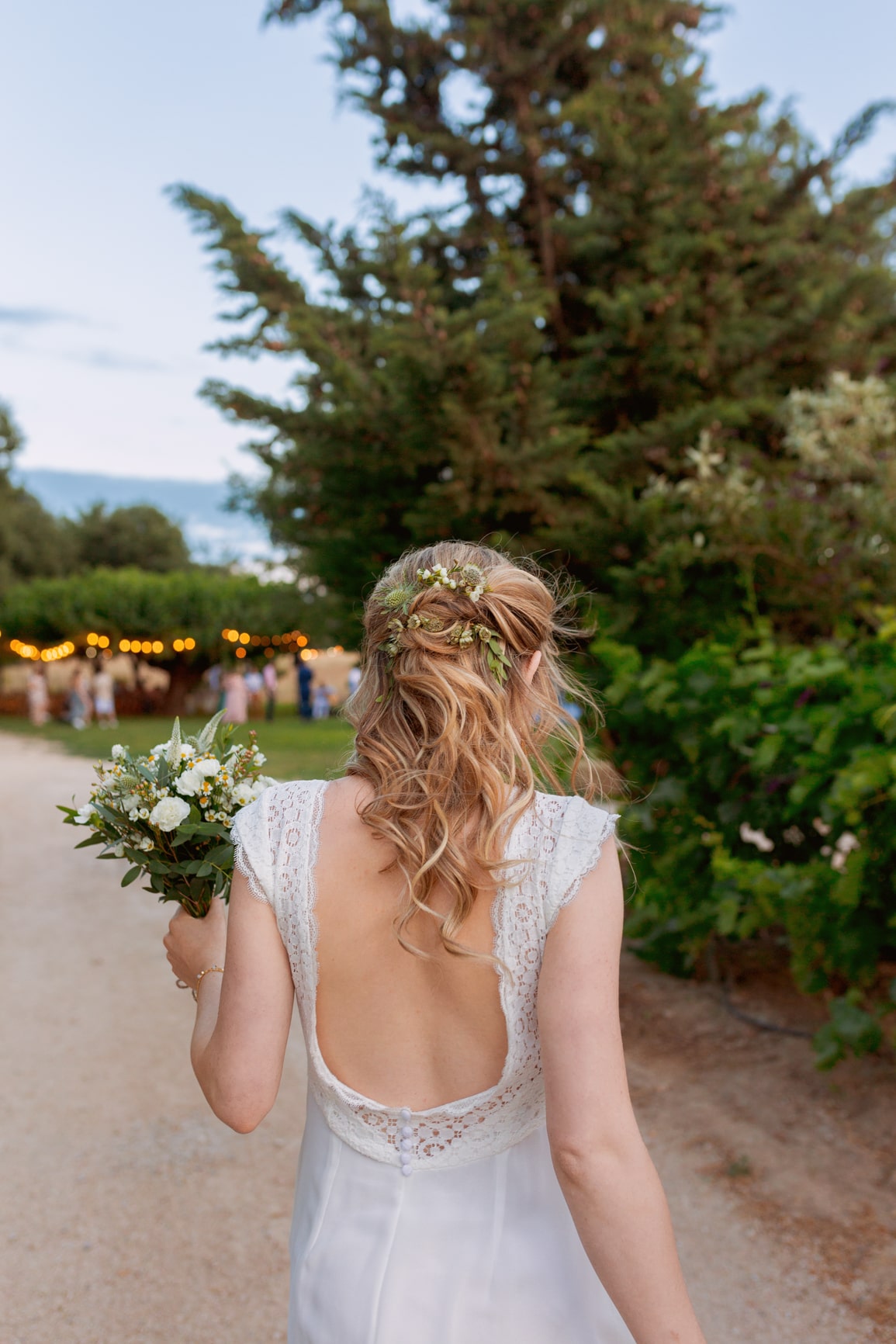 Vue de dos d’une mariée tenant son bouquet, marchant vers la réception.