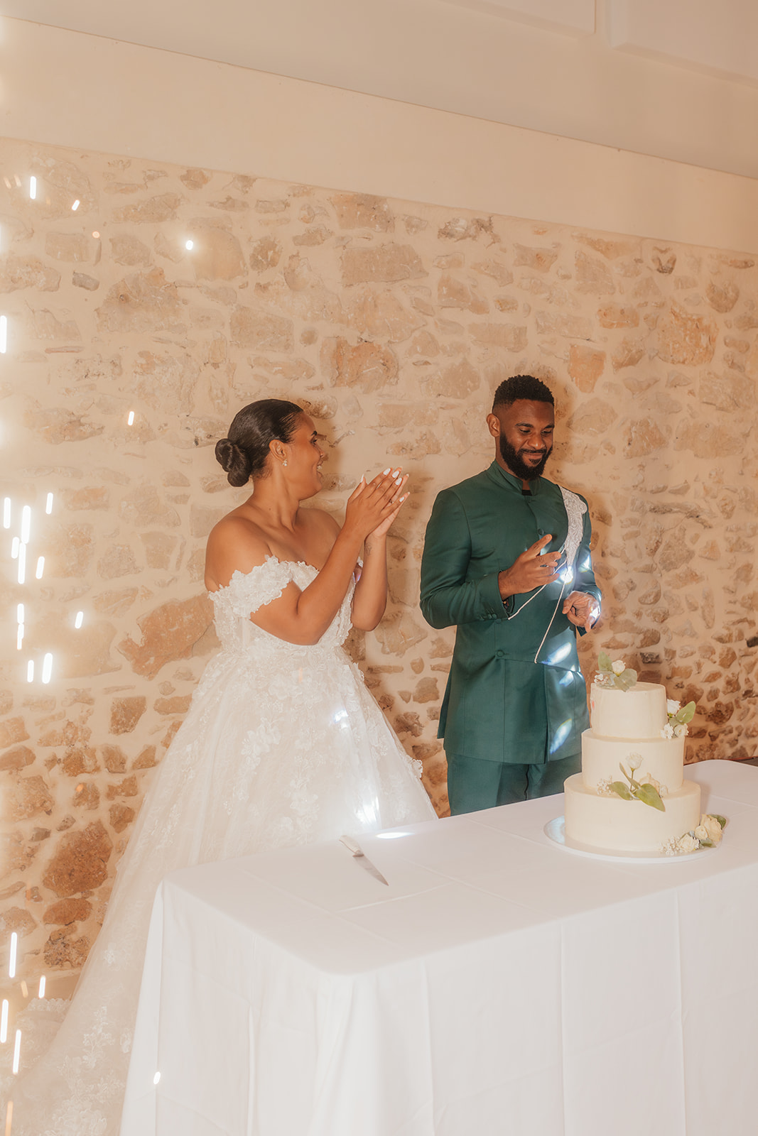 Les mariés rient devant leur gâteau de mariage, dans une salle en pierre lumineuse.