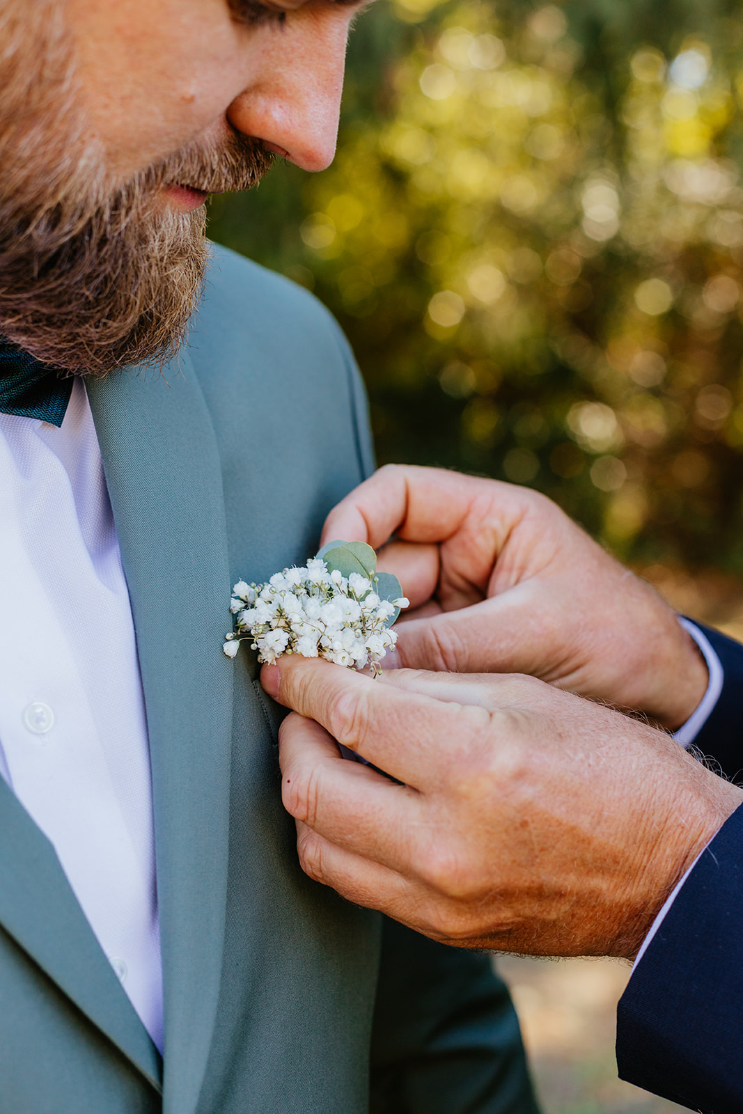 Un homme ajuste la boutonnière d’un marié portant un costume vert, lors des préparatifs de mariage.